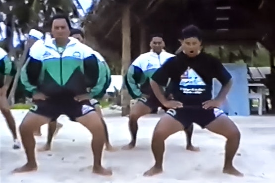 Several men are mid-haka or ceremonial dance on a sandy beach. The image is blurry as it is from old footage.