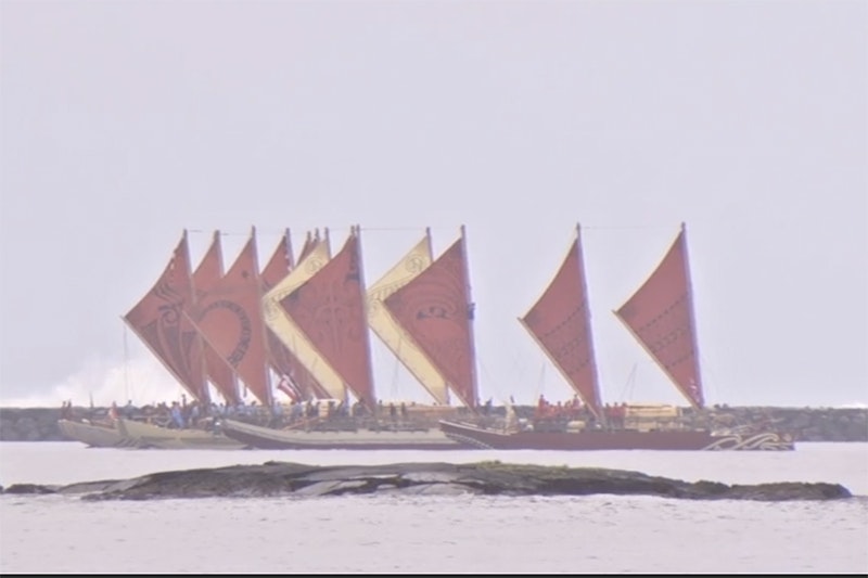 Several double-hulled waka with red and yellow sails are on the ocean.