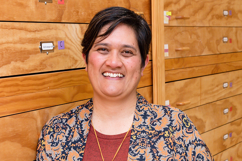 Head and shoulders photo of a woman leaning against wooden drawers and smiling.