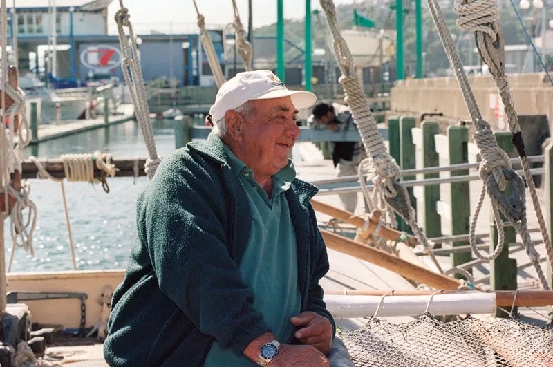An older man is sitting on a boat in a harbour and leaning against the railing.