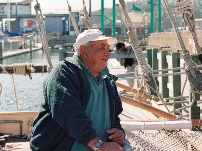 Te Aurere visit to Te Papa, 2000. Photo by Michael Hall. Te Papa (MA_I.036657) An older man is sitting on a boat in a harbour and leaning against the railing.
