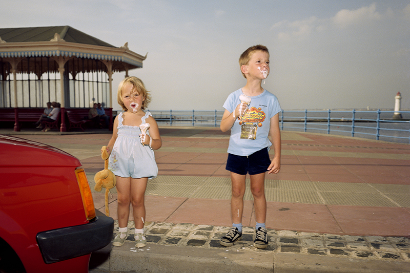 Two young children holding melting ice creams and with ice cream all over their mouths are standing on the side of the road next to a pier.