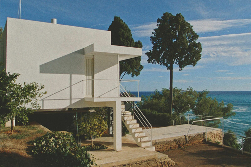 A moder boxy building with two trees in front of it is on a seaside cliff.