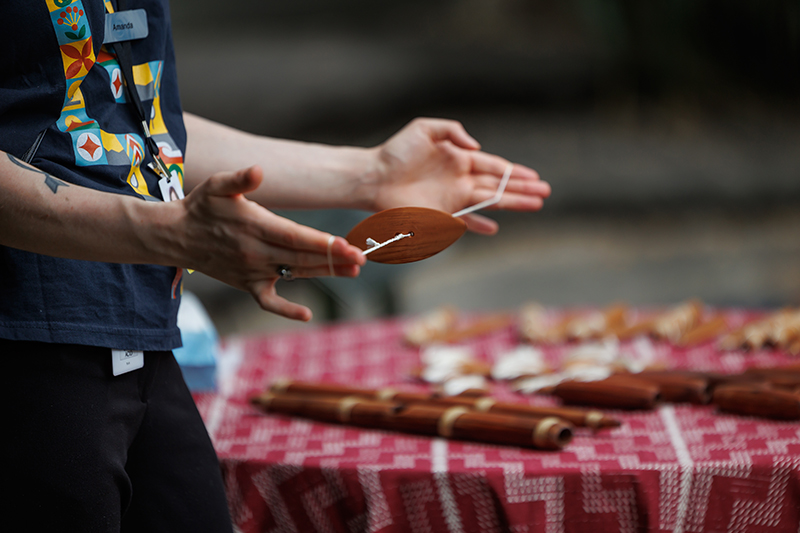 A pair of hands holding a piece of carved wood on a string between both hands. In the background is a table of wooden Māori instruments on a red cloth.