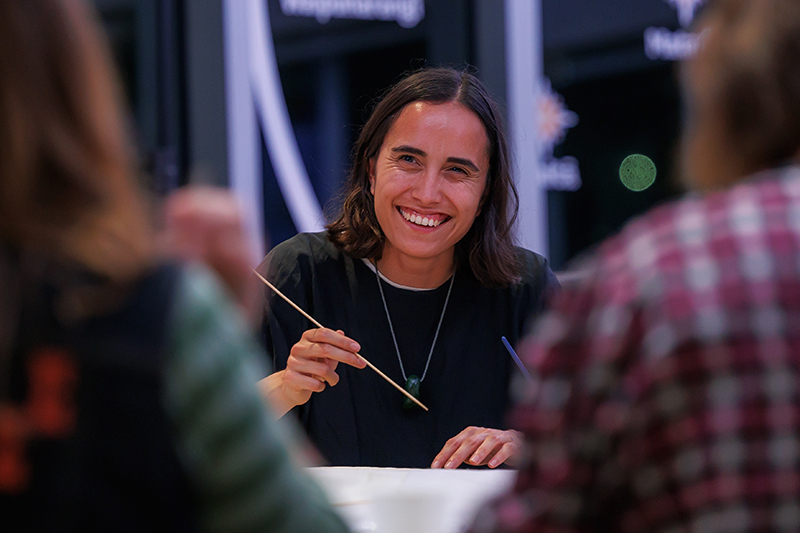 A woman is painting on white paper and smiling to someone opposite her.