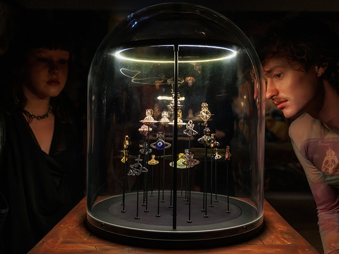 Vivienne Westwood & Jewellery, Te Papa, 2025 Two people are in a dark room looking at several small items of jewellery under a glass dome.