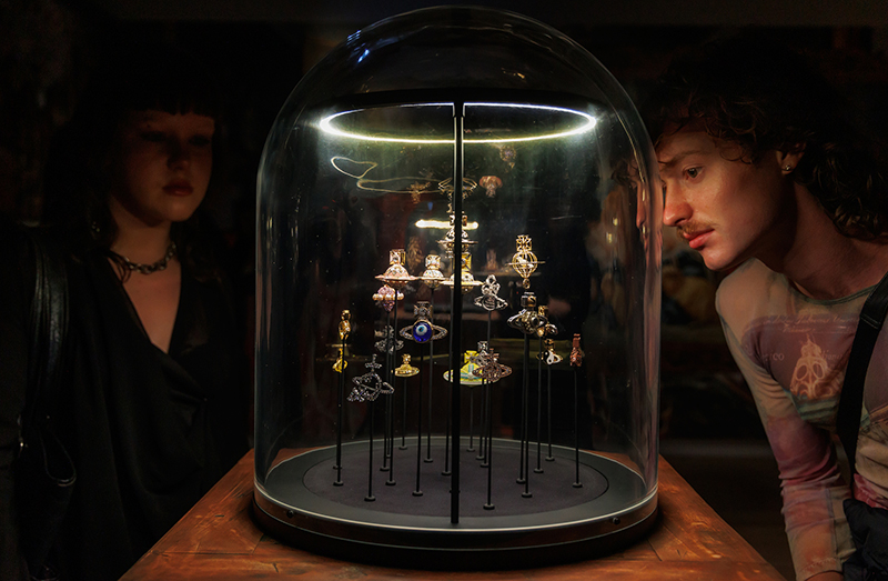 Two people are in a dark room looking at several small items of jewellery under a glass dome.