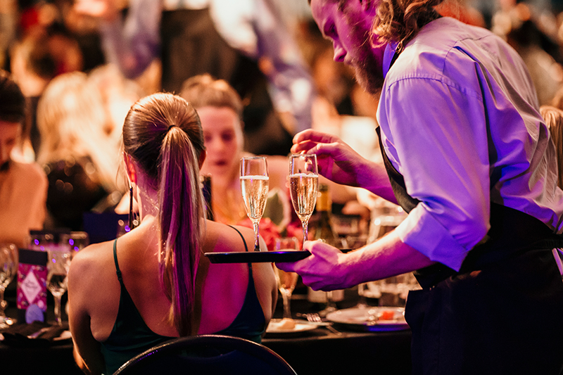 A waiter has a tray with two glasses of bubbles that he is handing to women sitting at a table.
