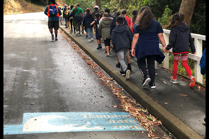 A group of primary school age children walking up a footpath next to a blue tsunami zone sign painted onto the road.