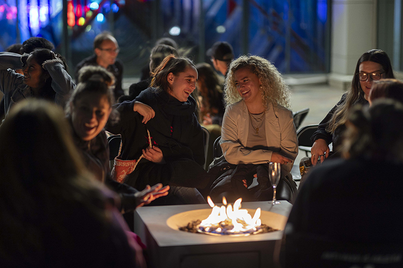 Several people sitting around a fire pit, laughing and talking  at night.