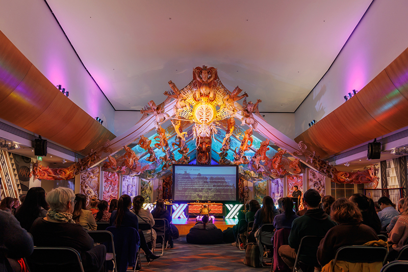 An audience watches a presentation beneath a carved colourful wharenui (meeting house).