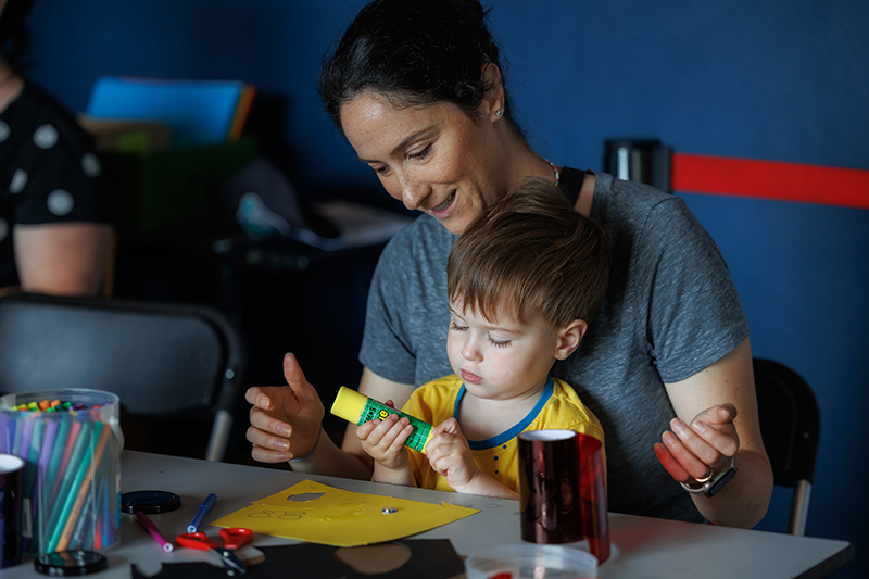 A woman has a child on her knee who is holding a tube of gluestick. They are making something on the table in front of them.
