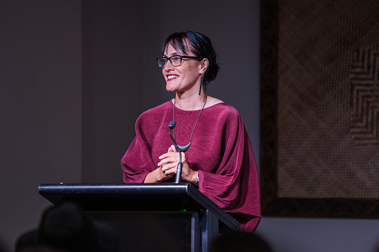 A woman wearing glasses and a red top is standing at a podium and smiling at people off camera.