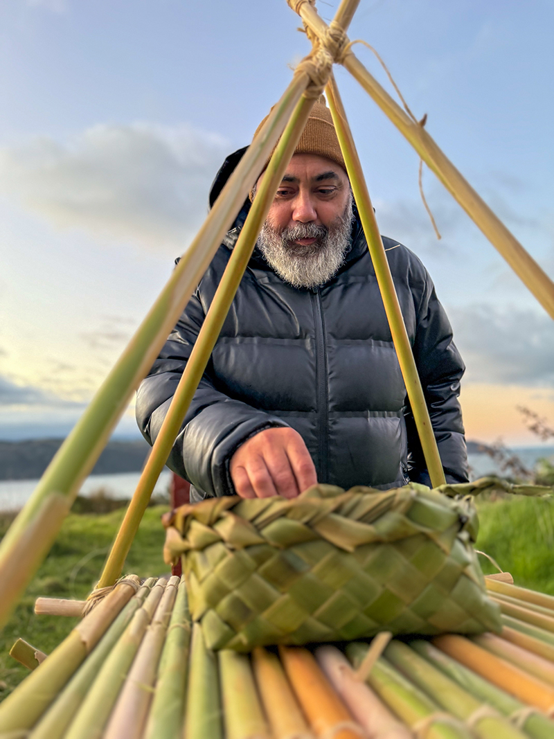 A man in a puffer jacket and beanie is reaching for something in a flax woven basket