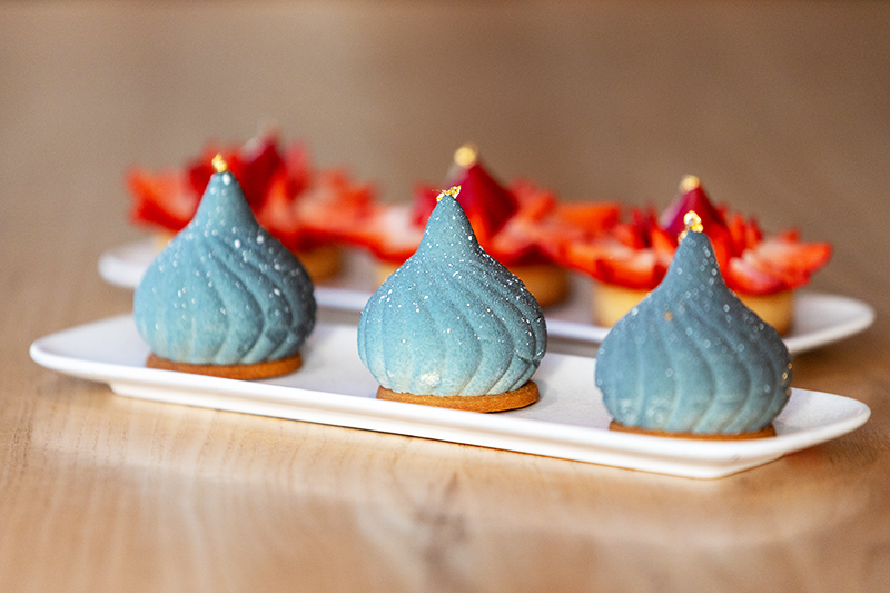 Two plates with two types of desserts arranged in three on each plate. The dessert in the foreground are three blue swirls with gold leaf on the top.