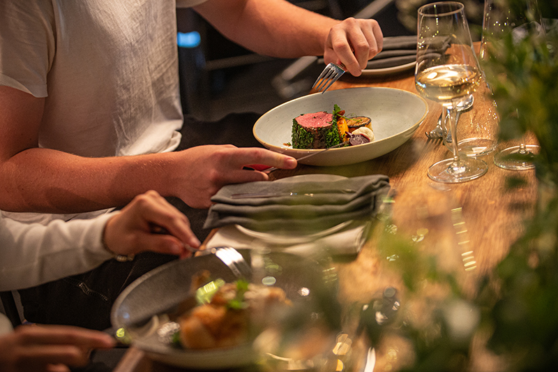 A person's arms and hands using a knife and fork to eat a meal on a table with plants and wine glasses.