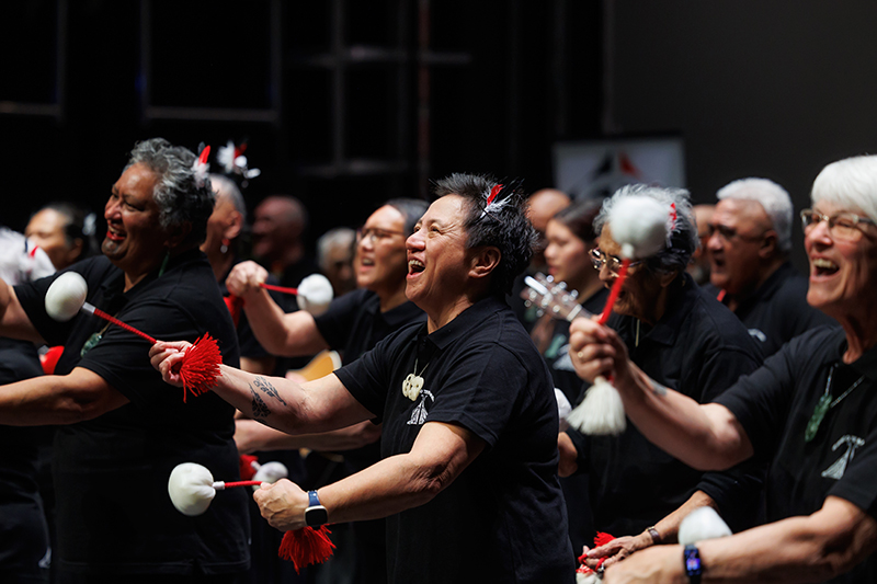 Several older people all wearing the same black shirt are singing and using Māori poi on stage.