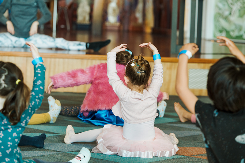 The backs of several children sitting on the ground with their arms in the air in a ballet pose. There is someone on the stage in the background showing them what to do.