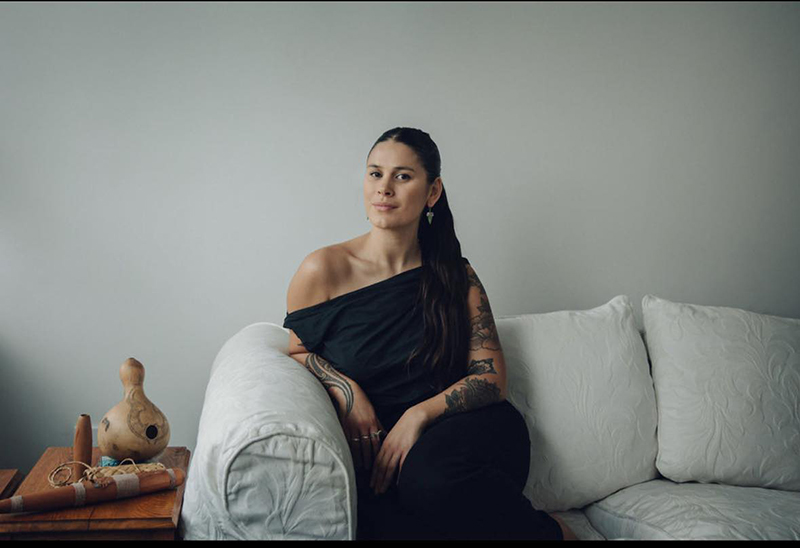 A woman wearing an off-shoulder dress is sitting on a cream couch. Next to her on a table are some Māori musical instruments.