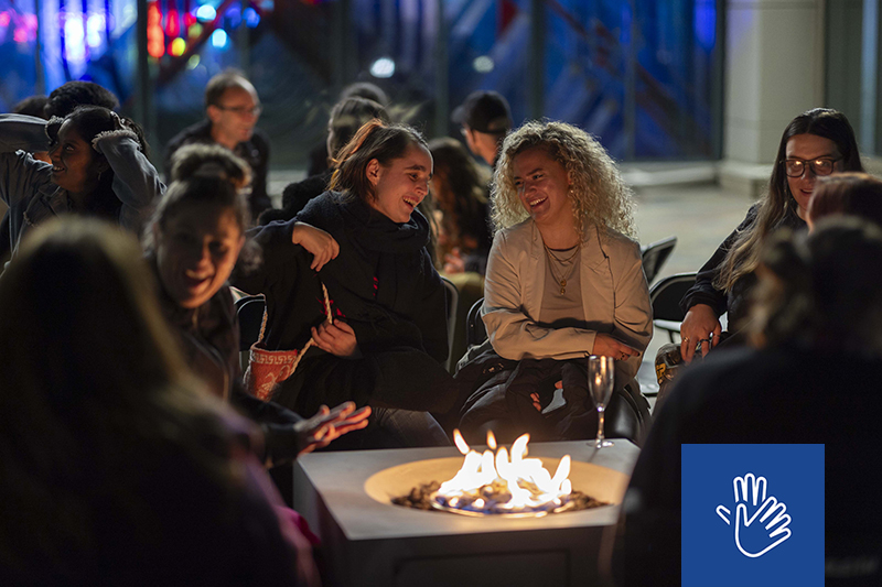 Several people sitting around a fire pit, laughing and talking  at night.