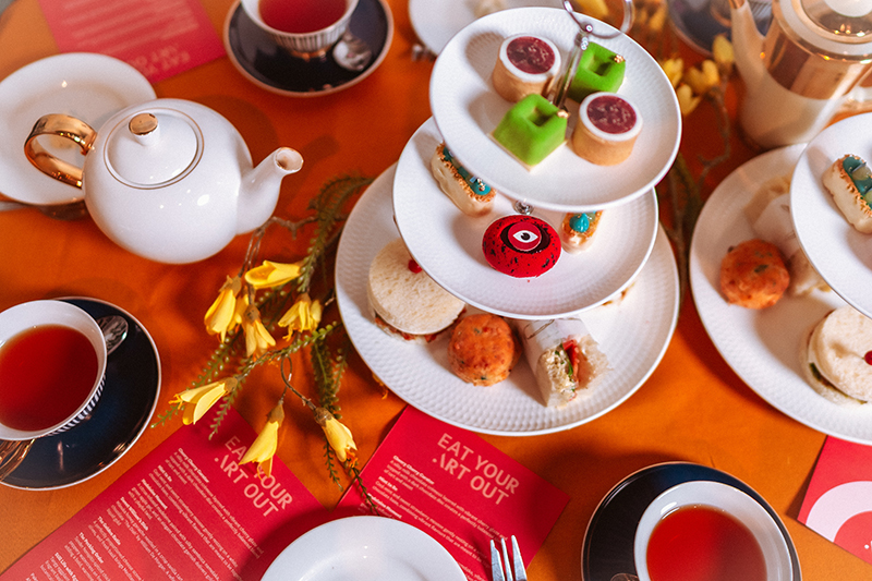 An image of an orange-red table with tea and teapots along with three-tierd plates with small cakes.