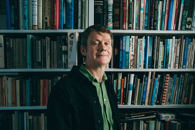 A man is standing three-quarters on in front of a bookshelf, looking at the camera.