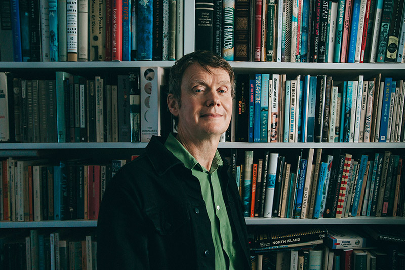 A man is standing three-quarters on in front of a bookshelf, looking at the camera.