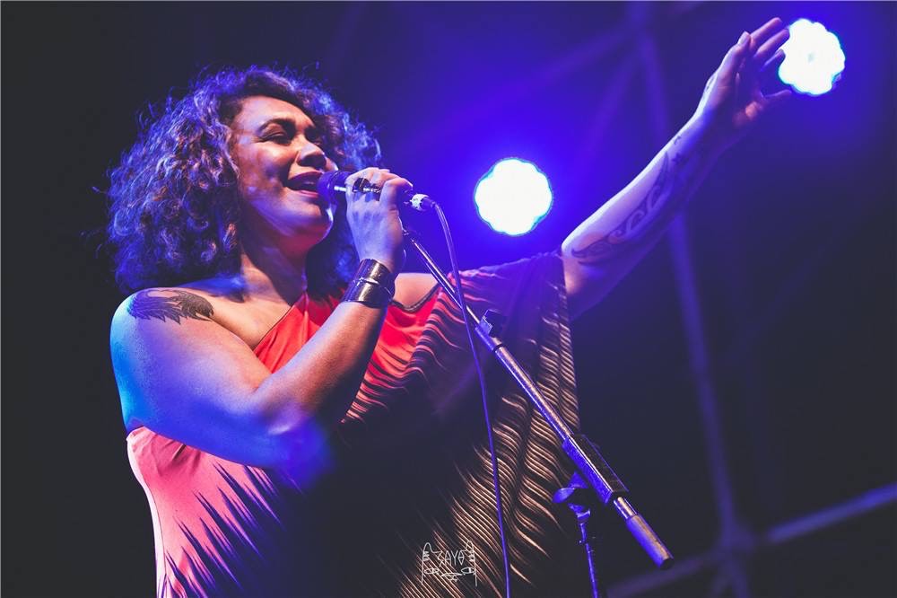 A woman in an off-shoulder outfit is singing into a microphone on a lit stage.