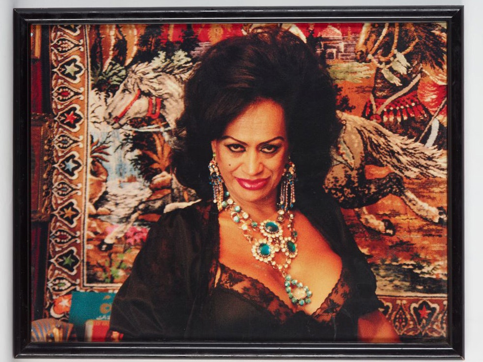 Carmen Rupe, by Jill Carter-Hansen, about 1986, Sydney. Purchased 2006. Te Papa (GH011926) A photo of a framed photograph of Carmen Rupe with big hair, a low-cut dress with large costume jewellery, and a carpet with horses running on it hung behind her.