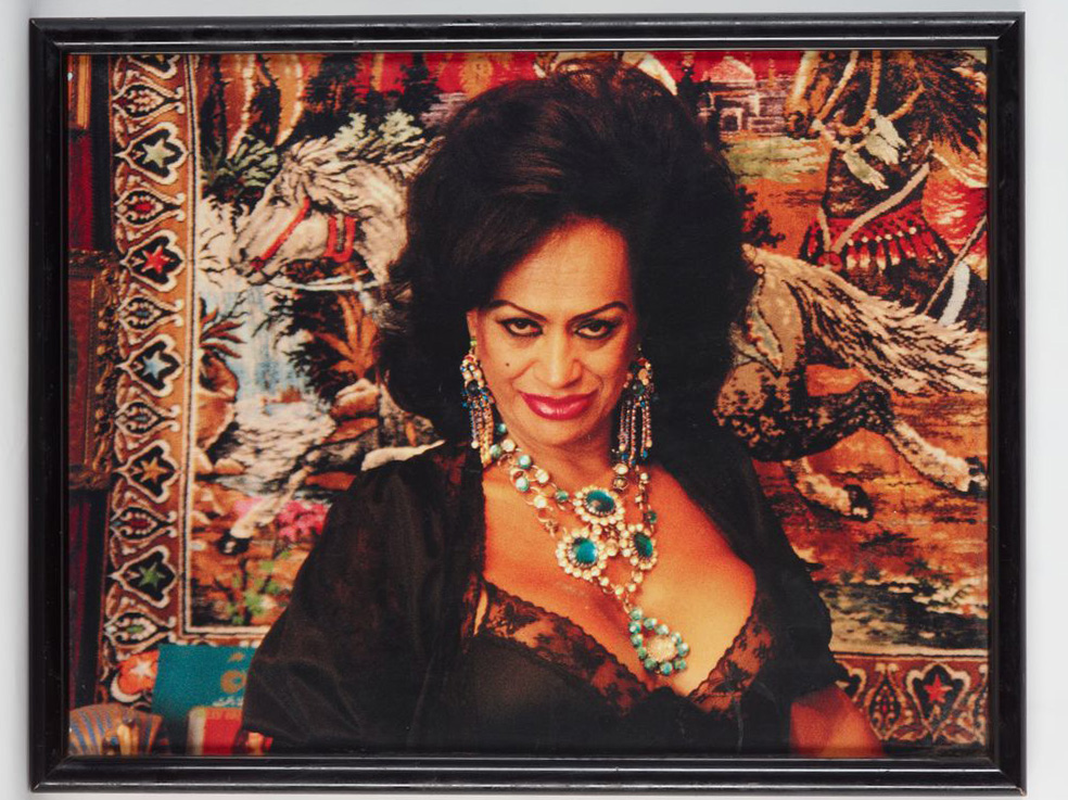 A photo of a framed photograph of Carmen Rupe with big hair, a low-cut dress with large costume jewellery, and a carpet with horses running on it hung behind her.