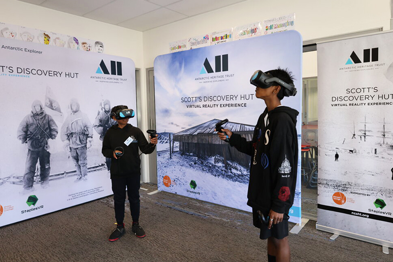 Two young boys wearng VR headsets and standing in front of three signs advertising an immersive antarctic virtual reality experience anotu Scott's Hut.. 