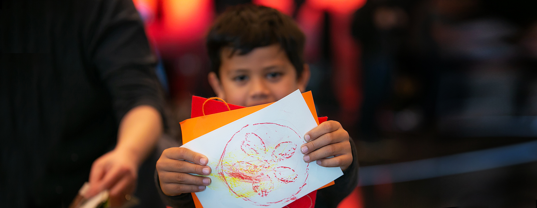A child is holding up a piece of paper with an image on it that they have either drawn or coloured in.