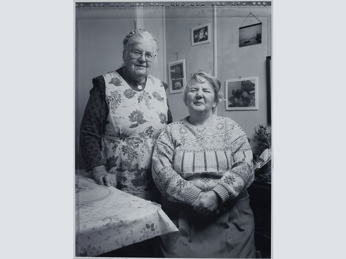 Anne Noble, Hidden Lives: the work of care. Gladys Thetford and her daughter Audrey, 1992-1994; Wellington. Purchased 2003. Te Papa ( O.027961/9) A black and white photo of two older woman smiling at the camera. One is standing by another sitting down.