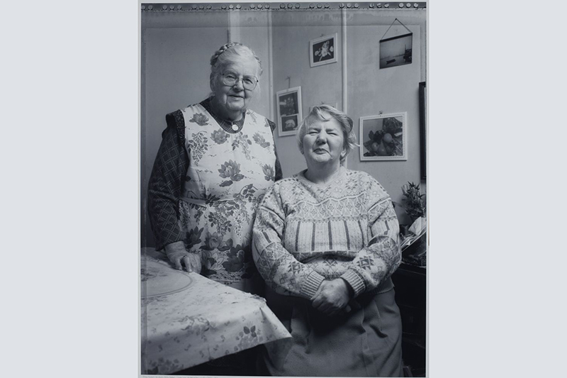 A black and white photo of two older woman smiling at the camera. One is standing by another sitting down.