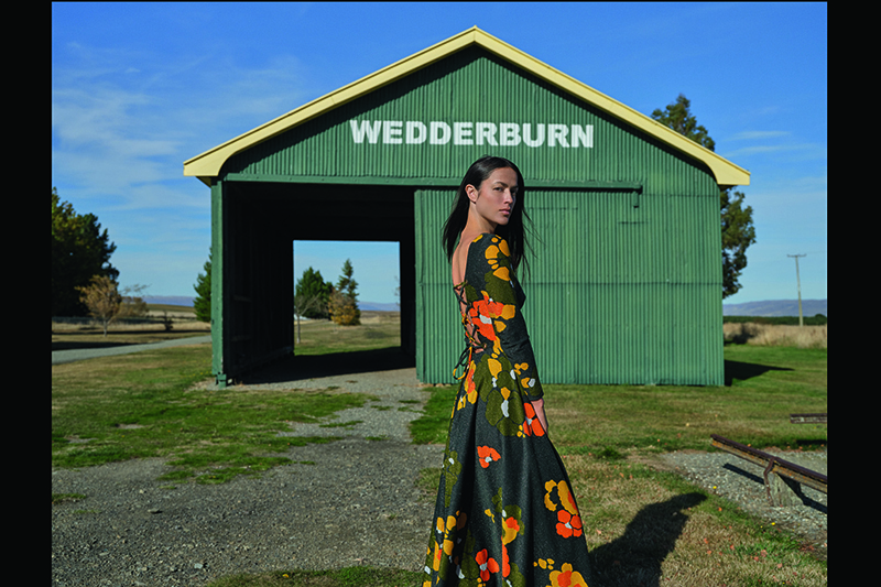 A photo of a woman standing in front of a green shed with the word Wedderburn painted on it. She is wearing a longsleeved long dress that is green with yellow and orange flowers on it.