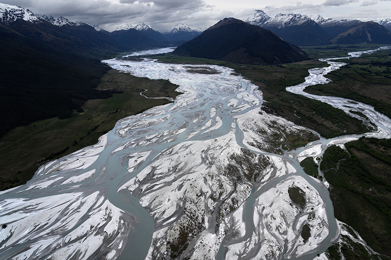 An aerial photo of a braided river with snow showing on the land on the river as well as the mountains in the far distance.