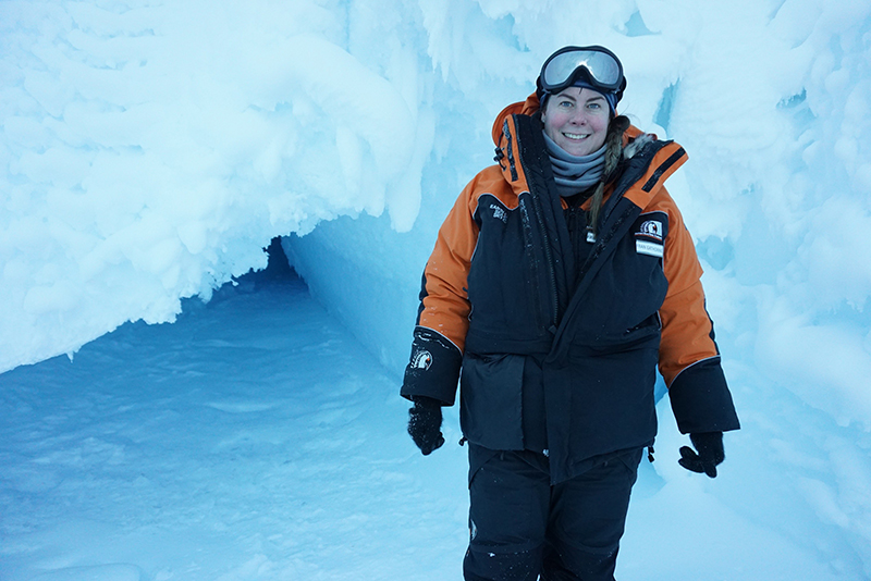 A woman in cold protective clothing and ski goggles on her head is standing in front of an ice cave and smiling at the camera.