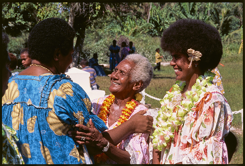 Three women in brightly coloured dresses are greeting each other on a Pacific Island.