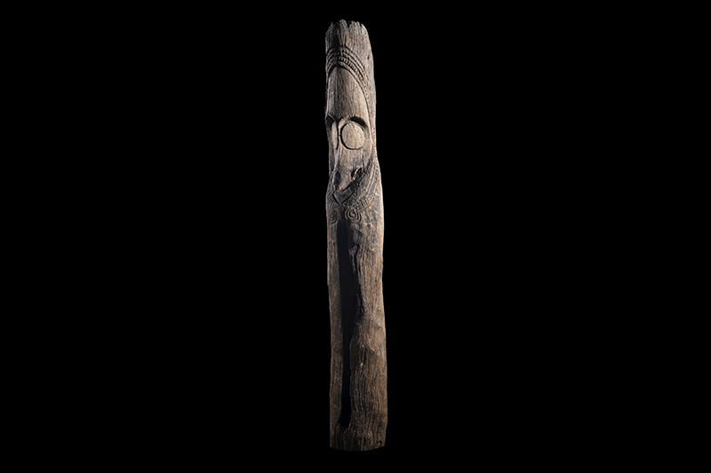 A tall carved wooden pole with a face in the top half and a vertical slit in the hollow bottom half.