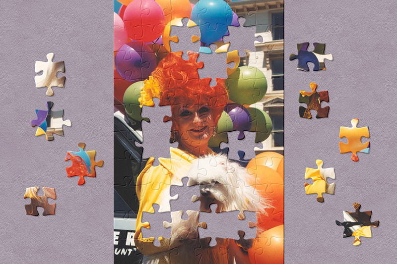 Jigsaw of a drag artist in a yellow gown cuddling a smiling, fluffy white dog. Their curly, red beehive wig is adorned with a yellow chrysanthemum hair piece. Rainbow balloons are attached to a car behind them.