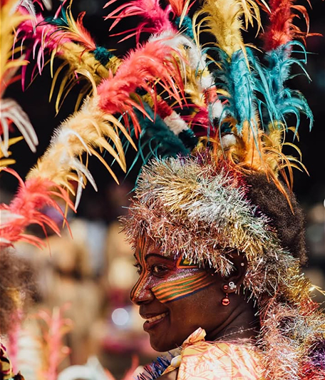 Vanuatu woman with her face painted with tribal motifs looking at the camera wearing a colourful head piece with colourful feathers and sparkly tinsel.  