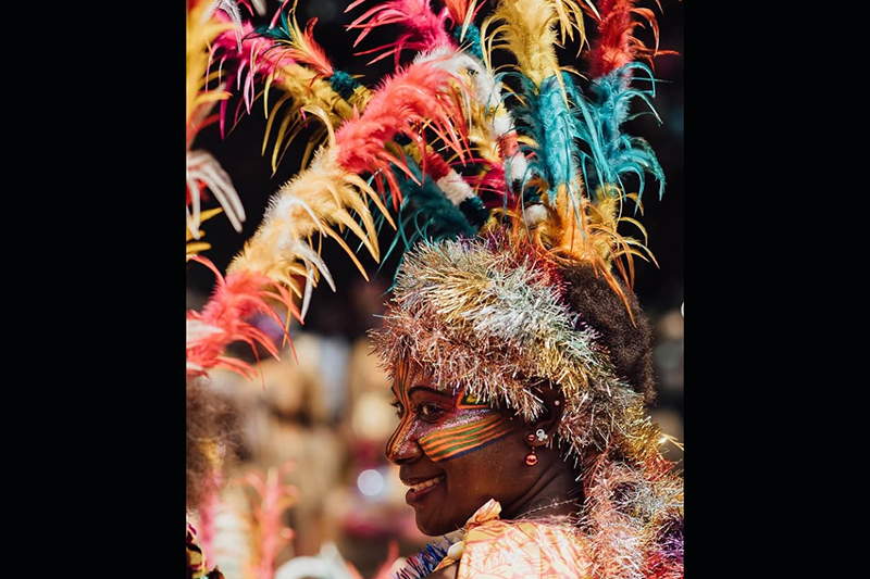 Vanuatu woman with her face painted with tribal motifs looking at the camera wearing a colourful head piece with colourful feathers and sparkly tinsel.  