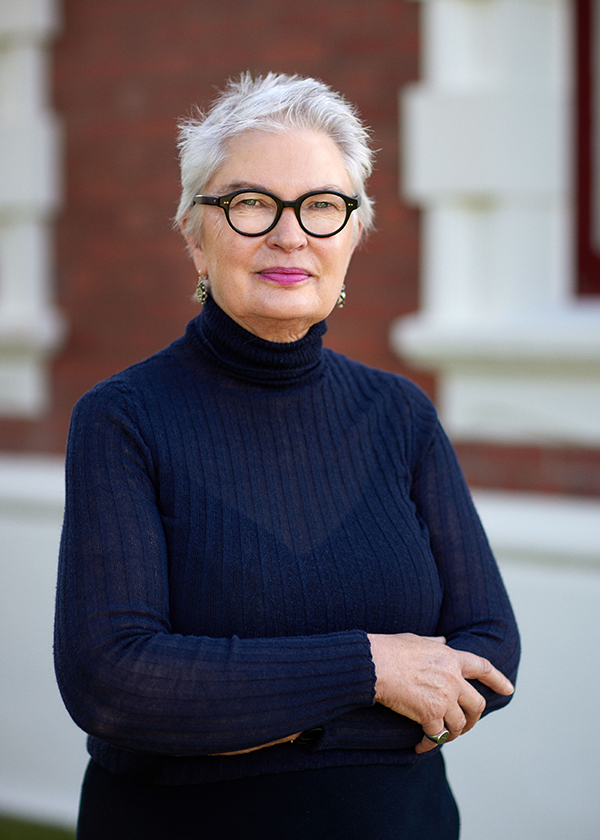 A woman with short grey hair, and black statement glasses has her arms crossed in front of her and is standing outside a brick building and looking at the camera.