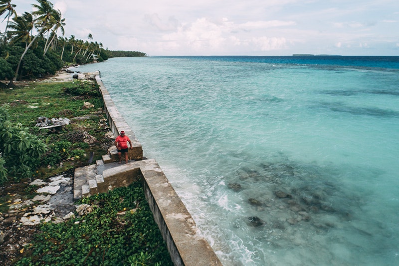 A man in a red tshirt is walking along a stone wall that divides a Pacific island and the sea.