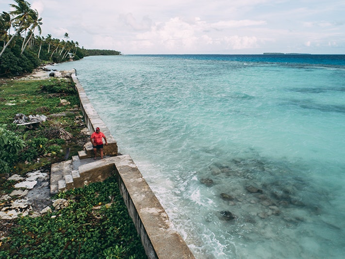 Photo by Andrew Matautia A man in a red tshirt is walking along a stone wall that divides a Pacific island and the sea.