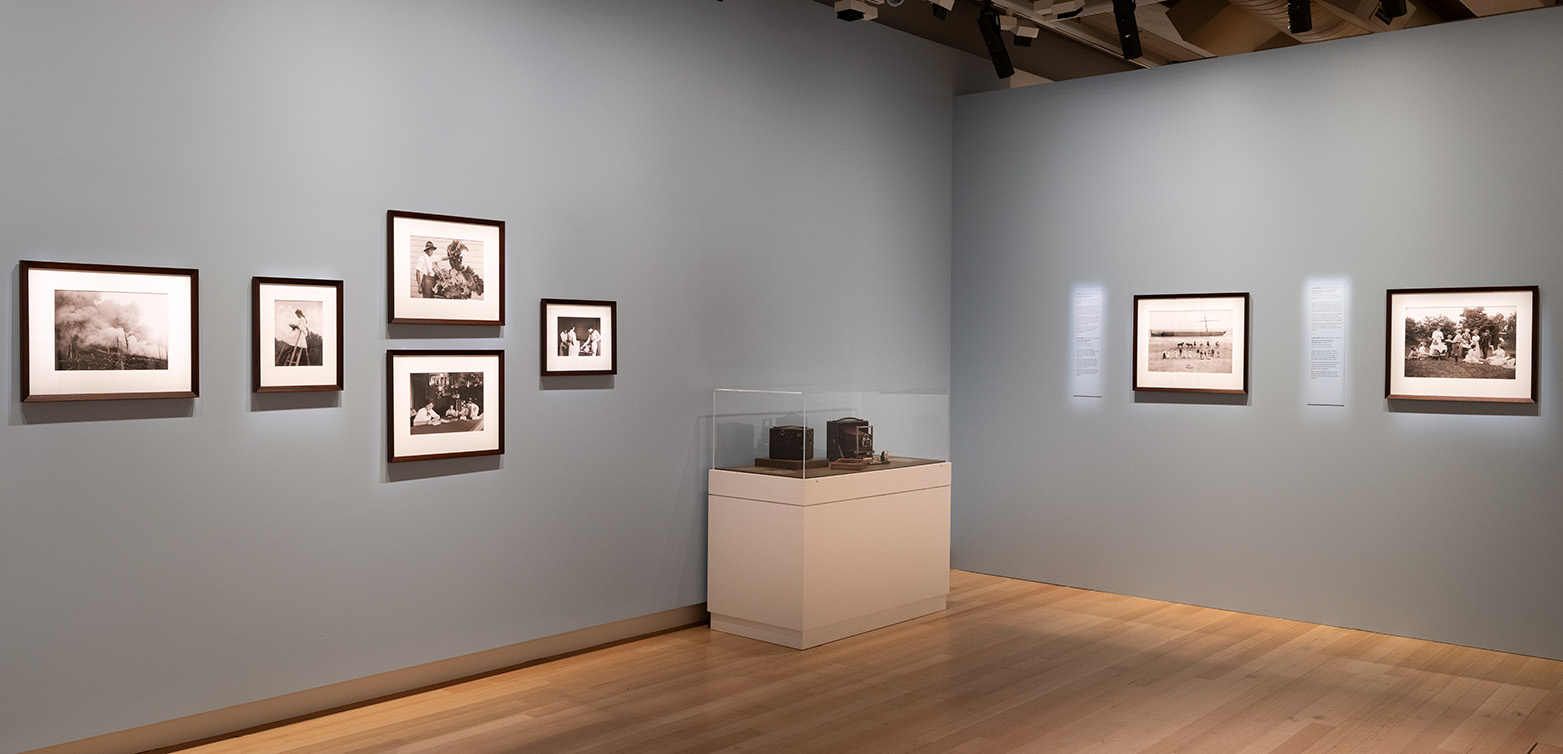 A museum gallery corner wall that is grey with seven framed photographs on the walls and a small cabinet of two box cameras.