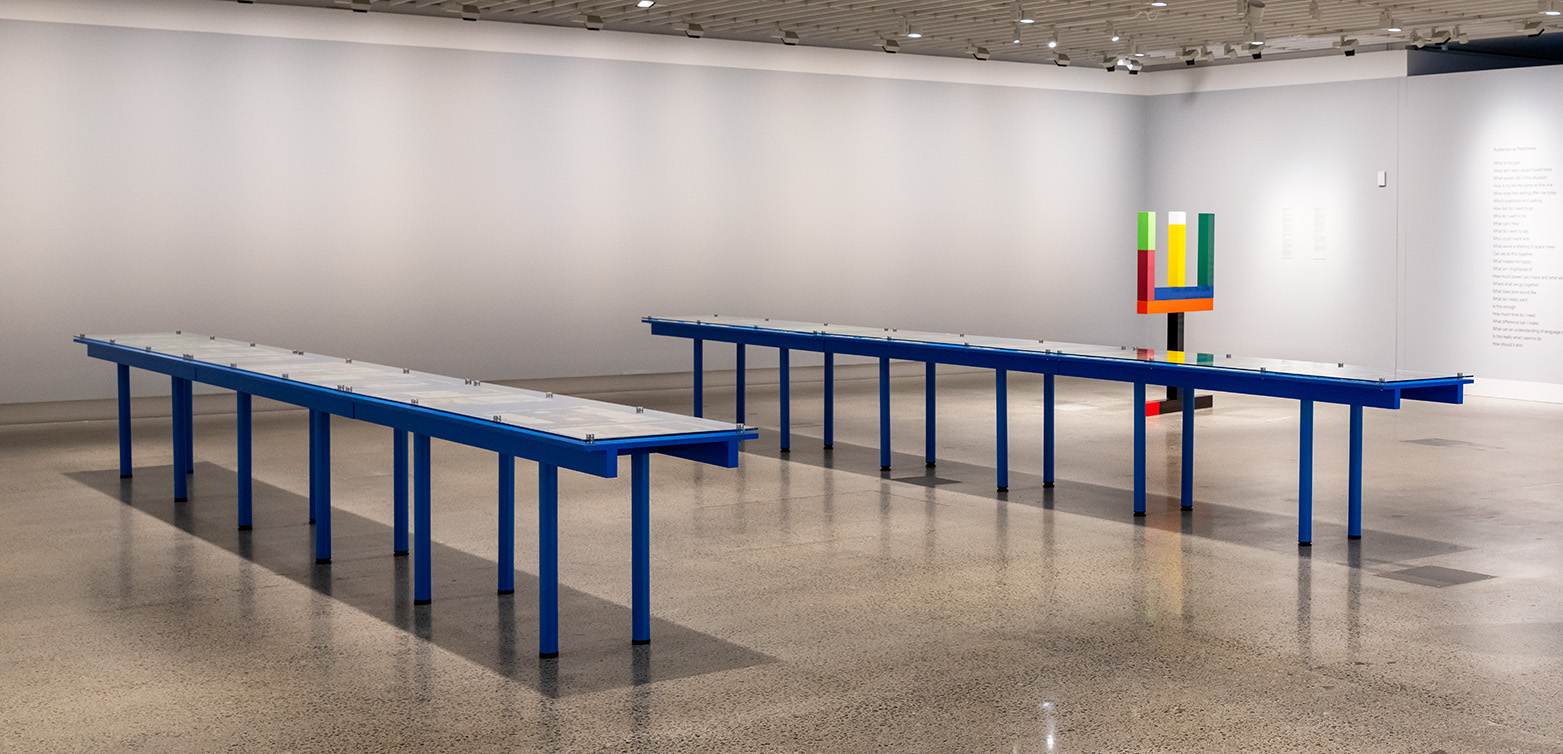 A large gallery room with a colourful sculpture in the background and two long tables with glass tops in the foreground. There are newspaper pages under the glass that are not visible in the photo.