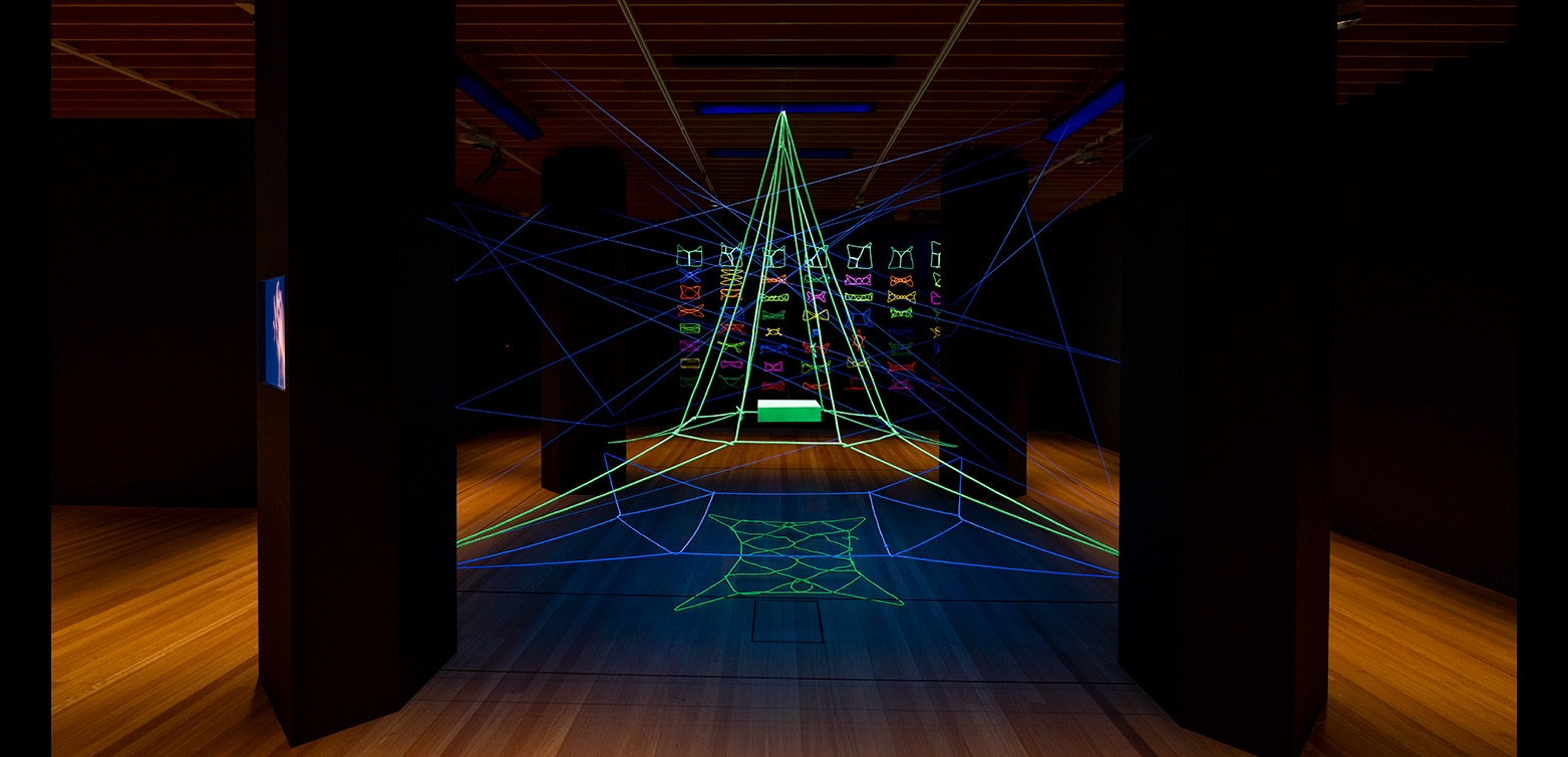 A room with four black pillars and some ultraviolet-lit string in different colours strung up between the pillars and on the wall behind it.