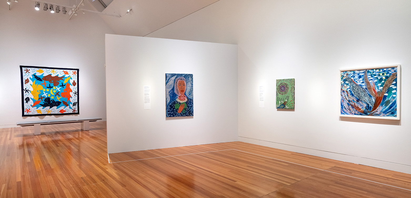 A gallery space with white walls and three paintings hung on the walls in the foreground, and a tīvaevae quilt hung on the back wall.