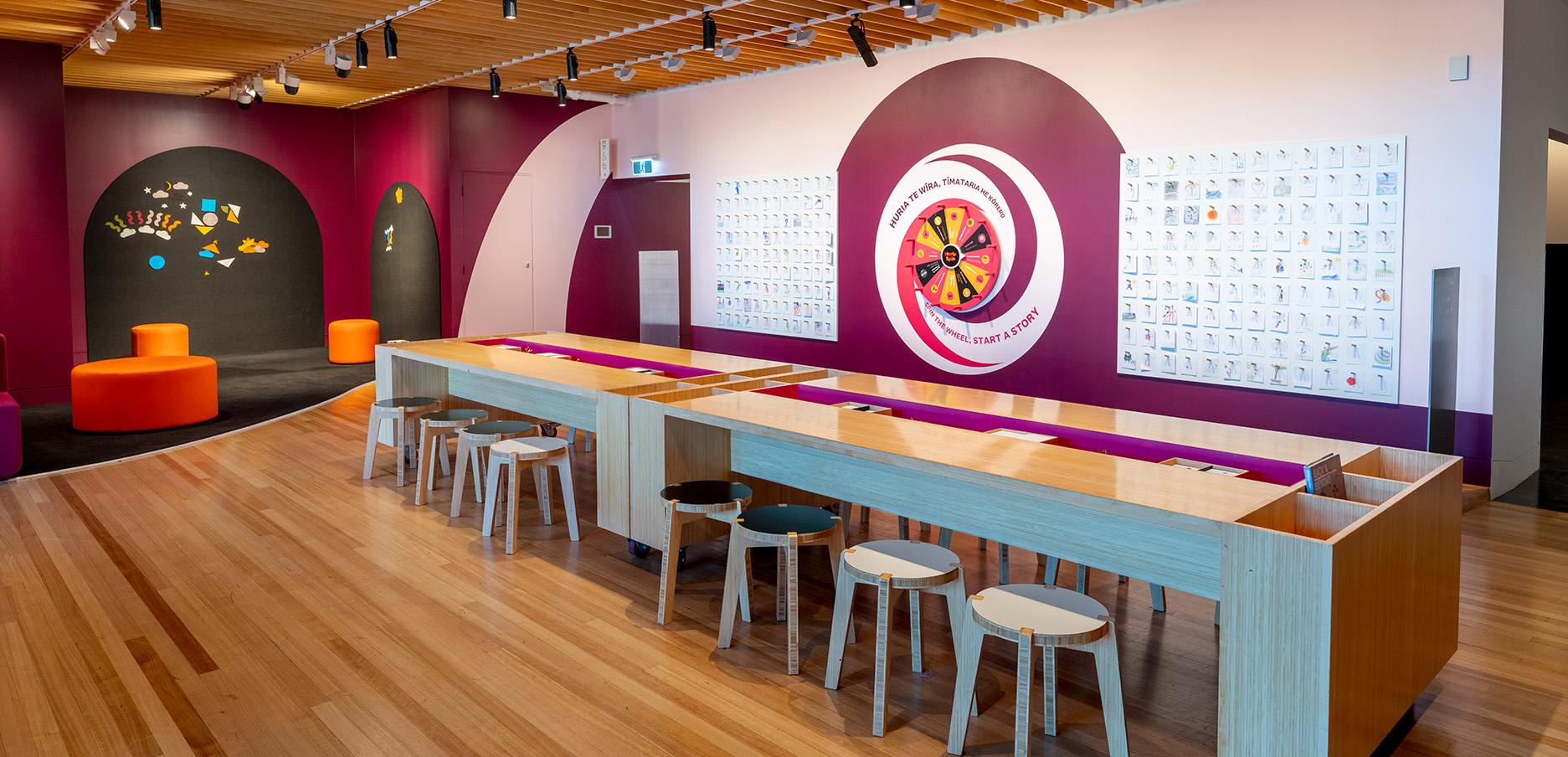 A room in a gallery where there is a long table with places to draw and a wall to post images that have been coloured in, and a wall at the back to hang pieces of felt.